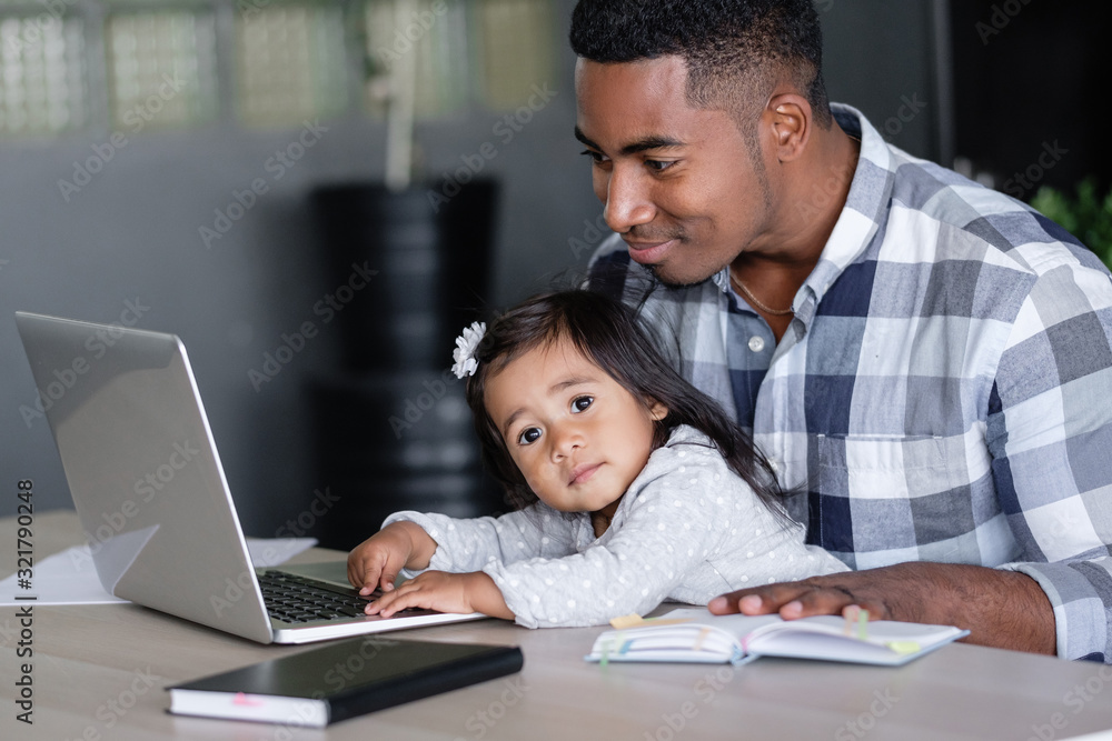 Young caring positive african-american father holding charming mixed race daughter in his arms pushing buttons of laptop while sitting at table with notebooks. Concept of combining business and family Young caring positive african-american father holding charming mixed race daughter in his arms pushing buttons of laptop while sitting at table with notebooks. Concept of combining business and family