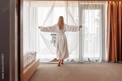 Joyful emotional young beautiful woman in white bathrobe happily opens curtains of her window in bedroom on sunny summer morning. The concept of starting a new day and fulfilling your plans