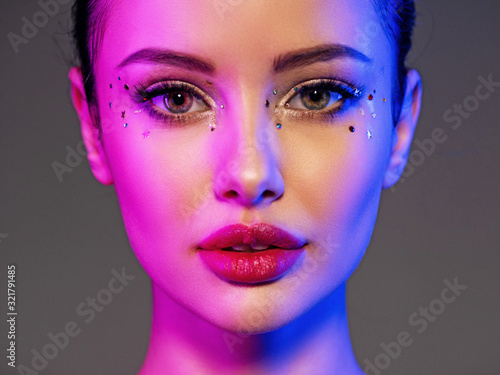 Portrait of the girl is highlighted in bright blue and purple light. Face of a beautiful model. Colorful portrait of a girl with bright makeup. Shiny sequins on the face. Art portrait. Sexy woman.