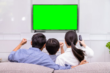 Back view of happy Asian family watching TV together in living room at home; cheerful and fun with movie, with green screen