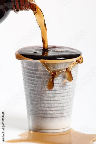 Pouring cola from bottle into overflowing plastic cup. Isolated on white background.