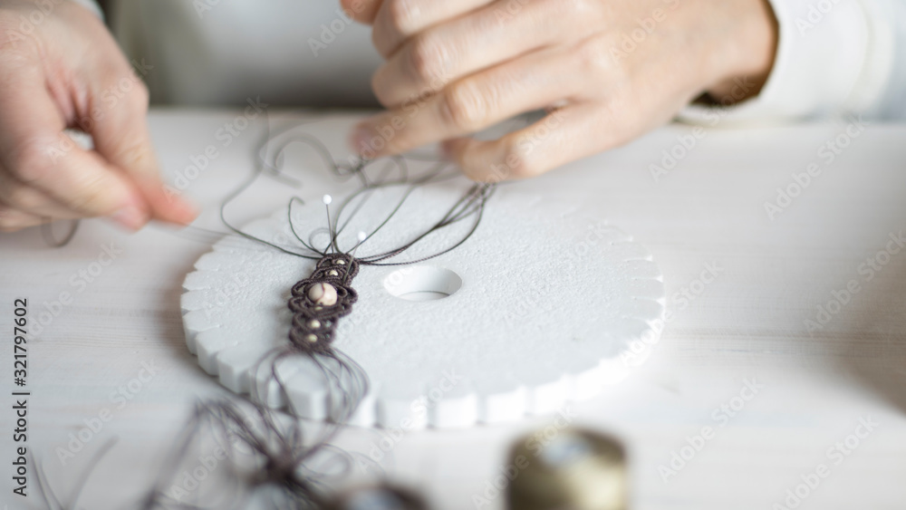 Obraz premium Lifestyle concept, work from home to reinvent your life: close-up of woman hands making macrame knotted jewelry with stone beads and tools on light wooden table