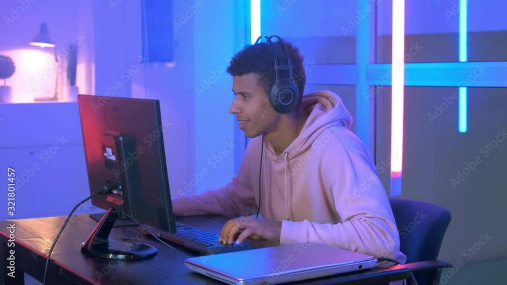 Medium shot of Persistent african gamer playing online shooter on pc in neon room. He spending his leisure time at home. Medium shot of Persistent african gamer playing online shooter on pc in neon room. He spending his leisure time at home.