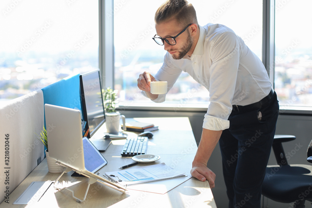 Confident young business man working on laptop and drinking from coffee cup while sitting in office. Confident young business man working on laptop and drinking from coffee cup while sitting in office.