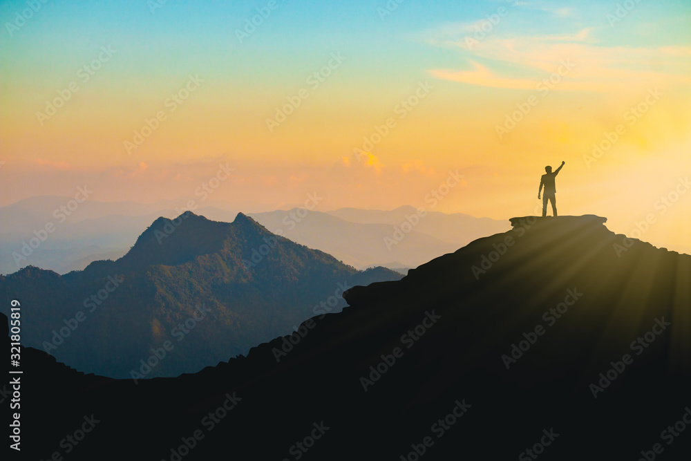 The silhouette of a man with a cheerful raise in his hand on the top of the mountain During sunset time The concept of effort and success