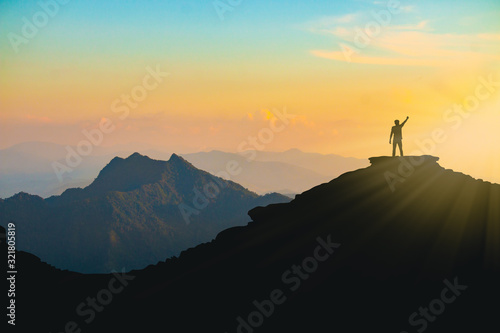 The silhouette of a man with a cheerful raise in his hand on the top of the mountain During sunset time The concept of effort and success © BNMK0819