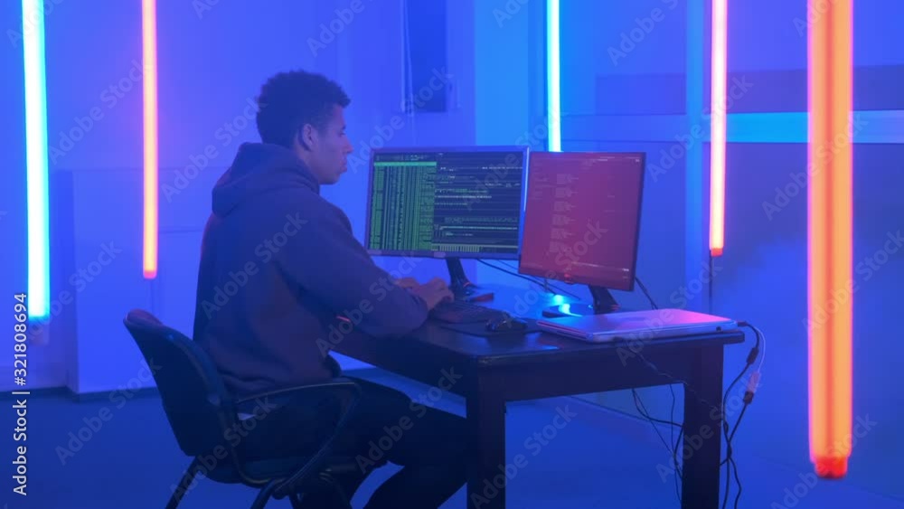 Young african man writing hacker programs in dark room with neon lights. He working on computer with two monitors. Young african man writing hacker programs in dark room with neon lights. He working on computer with two monitors.