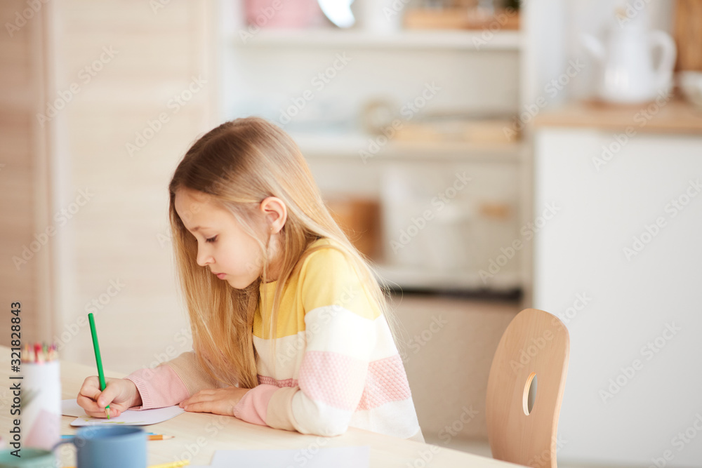 Side view portrait of cute little girl drawing pictures or doing homework while sitting at table in home interior, copy space Side view portrait of cute little girl drawing pictures or doing homework while sitting at table in home interior, copy space