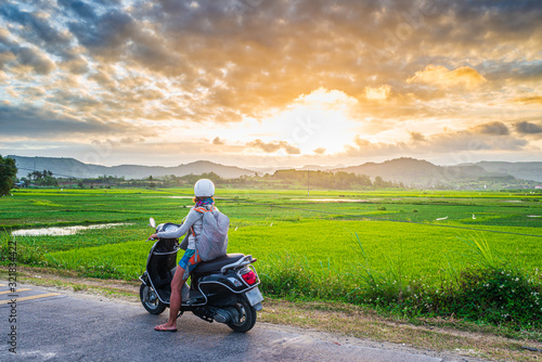 One person on motorbike looking at view of rice fields and mountains in the Phu Yen province, Nha Trang Quy Nhon, adventure traveling in Vietnam. Rear view sunburst backlight dramatic sky at sunset.