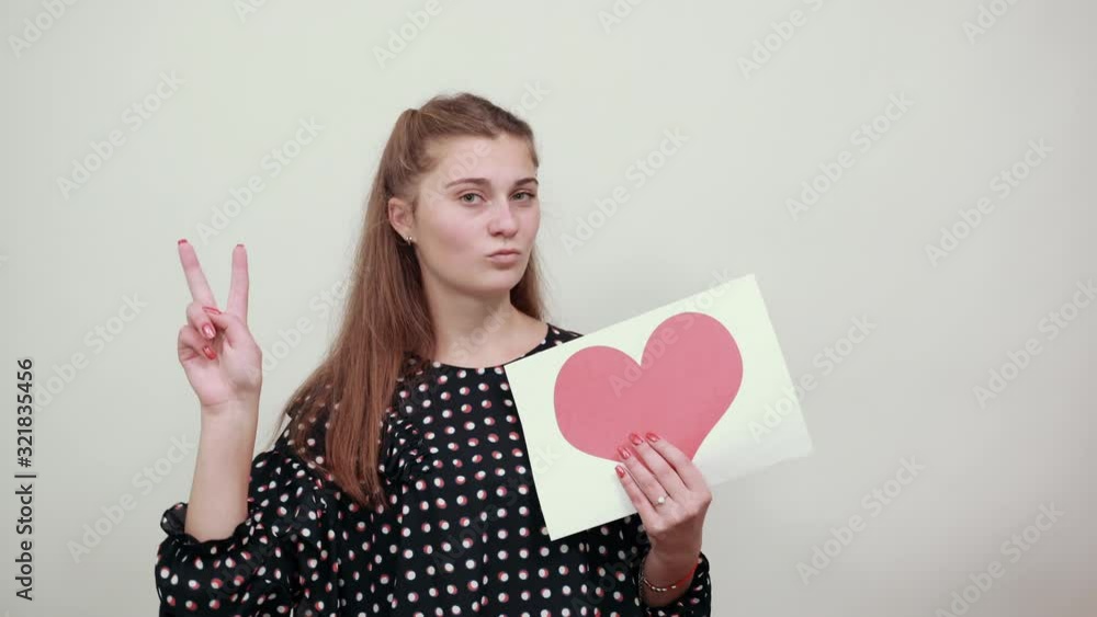 Fair-haired beautiful girl in a black dress with white circles holds piece of paper with heart shape and four fingers up Fair-haired beautiful girl in a black dress with white circles holds piece of paper with heart shape and four fingers up
