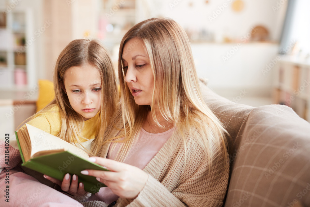 Warm-toned portrait of mature mother reading book to little girl while sitting on sofa in cozy home interior Warm-toned portrait of mature mother reading book to little girl while sitting on sofa in cozy home interior