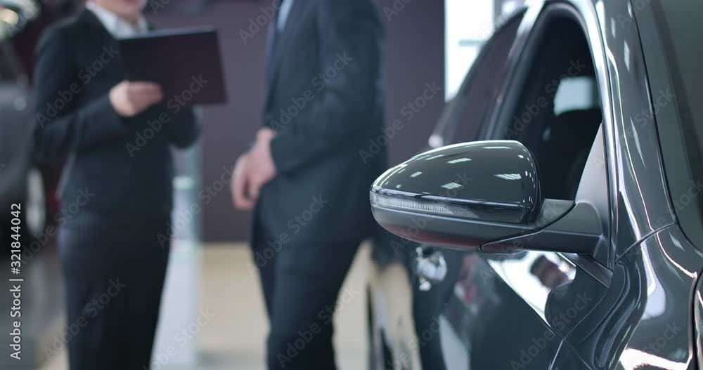 Unrecognizable Caucasian trader and client signing sale and purchase agreement in auto showroom. Focused on black car standing at the foreground. Selling, purchasing, industry. Cinema 4k ProRes HQ. Unrecognizable Caucasian trader and client signing sale and purchase agreement in auto showroom. Focused on black car standing at the foreground. Selling, purchasing, industry. Cinema 4k ProRes HQ.