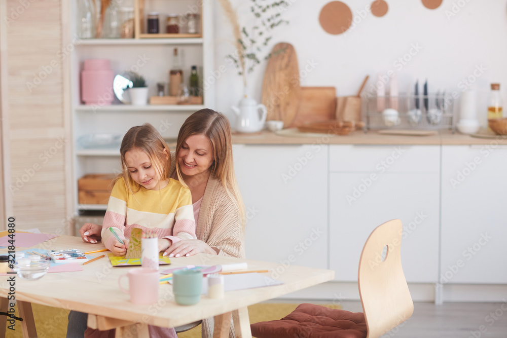Warm-toned portrait of happy mother hugging little daughter while painting pictures at wooden table in kitchen, copy space Warm-toned portrait of happy mother hugging little daughter while painting pictures at wooden table in kitchen, copy space