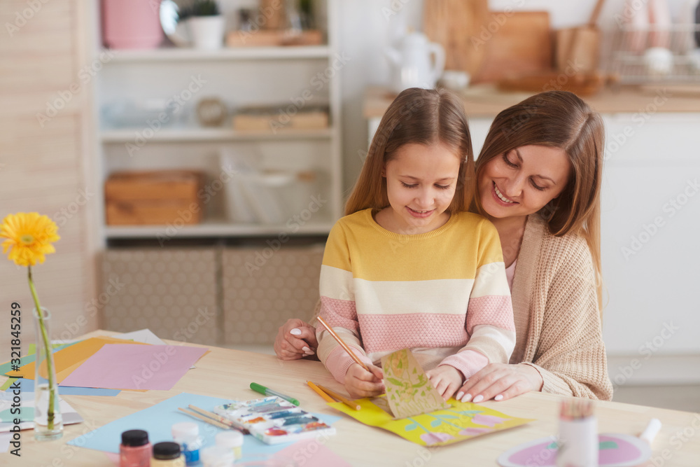 Fototapeta premium Warm-toned portrait of happy mother hugging daughter while drawing pictures at wooden kitchen table, copy space