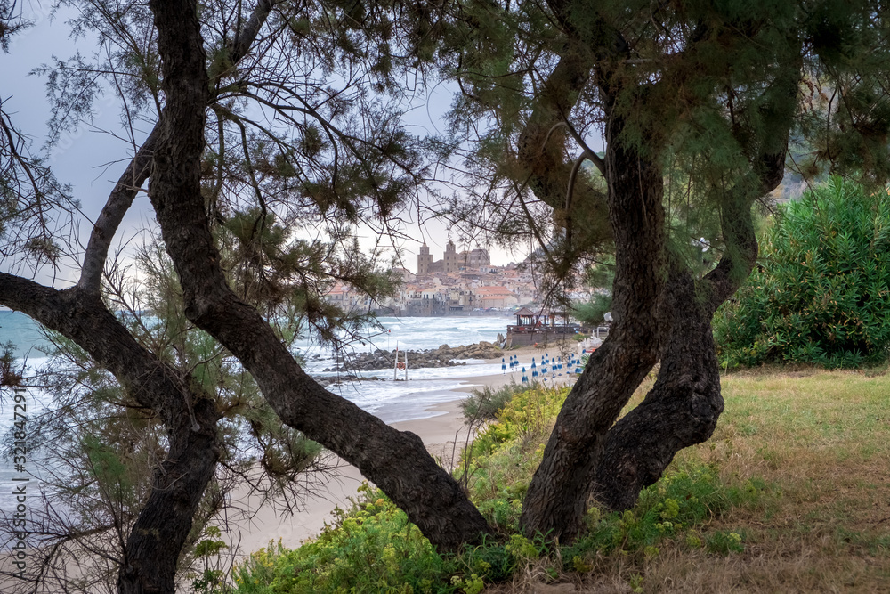 Fototapeta premium Seafront of Tyrrhenian Sea. View of the Sicilian city of Cefalu through trees growing on the shore