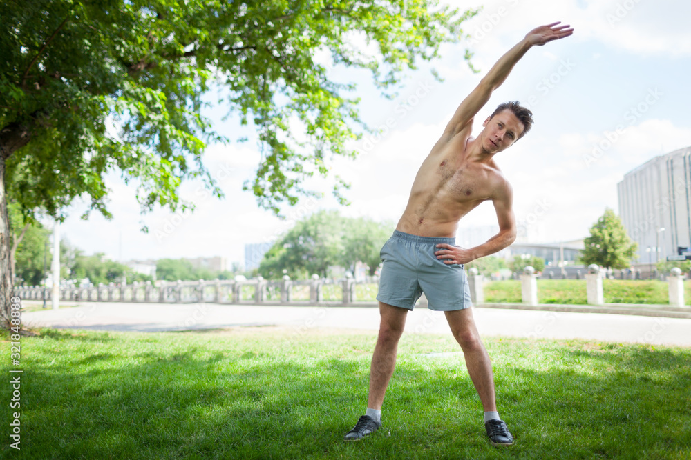 Front view of handsome gentleman with naked torso doing morning gymnastics outdoors. Sporty young man in shorts doing warm-up exercise on street. Concept of sport.