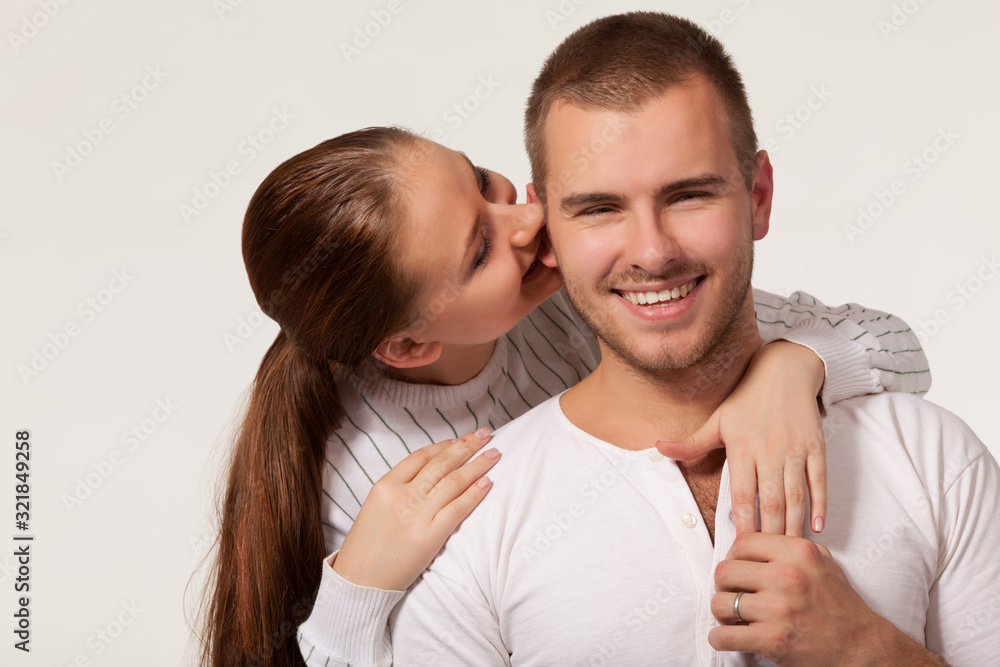 Front view of charming young woman hugging joyful guy from behind and biting ear. Handsome man holding girlfriend hand and smiling. Isolated on white studio background. Concept of relationships. Front view of charming young woman hugging joyful guy from behind and biting ear. Handsome man holding girlfriend hand and smiling. Isolated on white studio background. Concept of relationships.