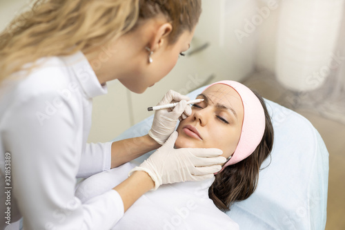Marking eyebrows with a pencil. Permanent makeup, tattooing of eyebrows. Cosmetologist in white gloves applying make up for woman in beauty salon