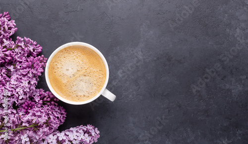 White cup with coffee and spring lilac flowers on black stone background. Copy space. Top view