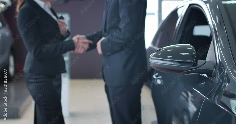 Unrecognizable Caucasian man and woman passing car keys and shaking hands at the background. Focused on black automobile standing at the foreground in dealership. Cinema 4k ProRes HQ. Unrecognizable Caucasian man and woman passing car keys and shaking hands at the background. Focused on black automobile standing at the foreground in dealership. Cinema 4k ProRes HQ.