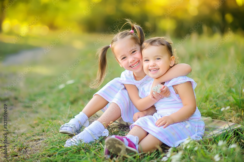 Cute children are walking in the park at sunset. Girls hug while sitting on the grass and laugh cheerfully. Children play and have fun outdoors. Cute children are walking in the park at sunset. Girls hug while sitting on the grass and laugh cheerfully. Children play and have fun outdoors.