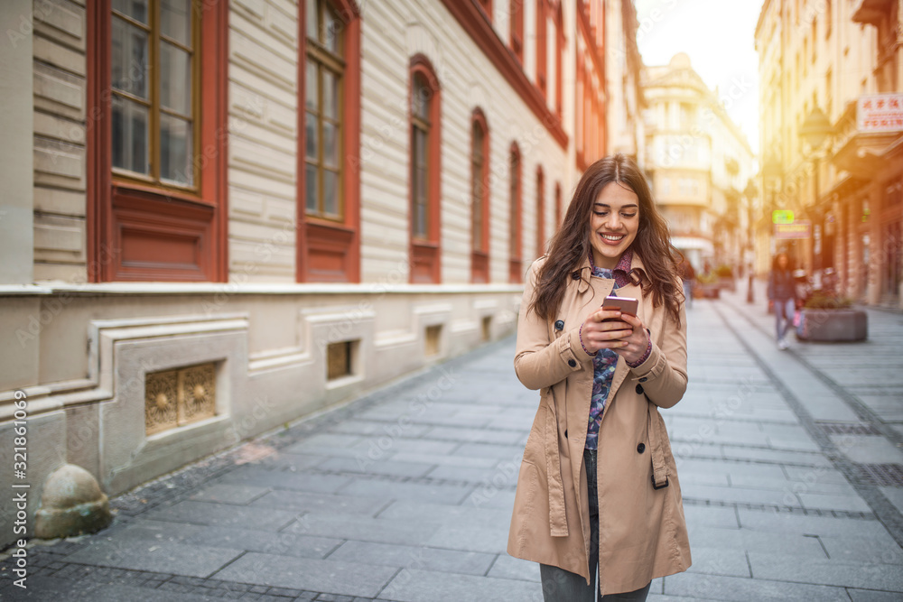 Fototapeta premium Photo of a woman using smart phone. Beautiful woman spending time in the city. Smiling young woman walking outdoors at urban setting and checking messages.