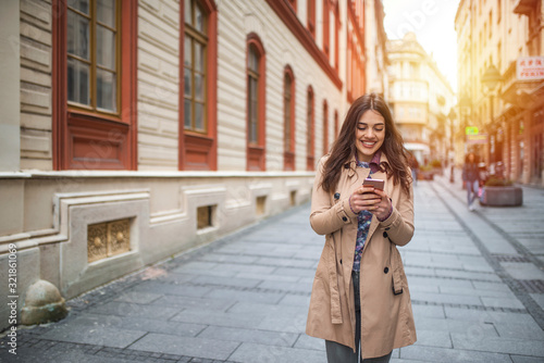 Photo of a woman using smart phone. Beautiful woman spending time in the city. Smiling young woman walking outdoors at urban setting and checking messages.