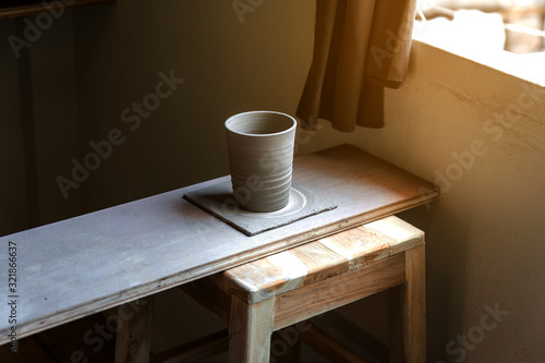 Hand crafted ceramic cup waiting for settling on wooden plate near the window. Creative workshop. 
