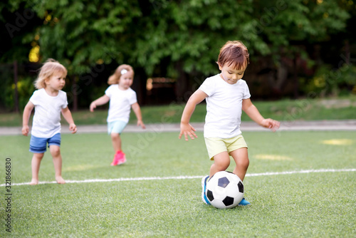 Football game: team of toddlers playing soccer on green field: three children, two boys (one is barefoot) and girl playing at stadium, smiling little boy dribbling ball is running in front place © AMR Studio