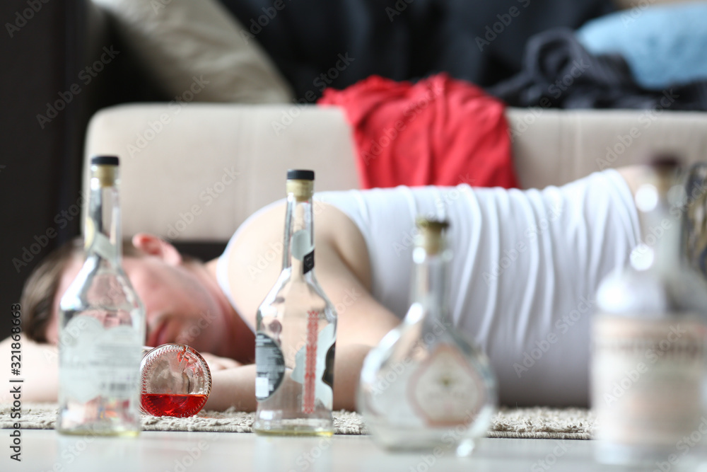 Empty bottles placed on floor man's apartment. Man lies on carpet near sofa with decorative pillows and sleeps after noisy evening. There is drink in front him, and things scattered behind.