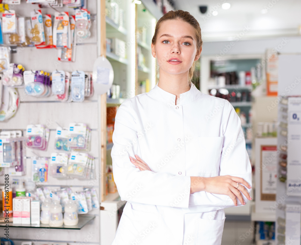 Cheerful woman pharmacist standing with arms crossed in interior of pharmacy