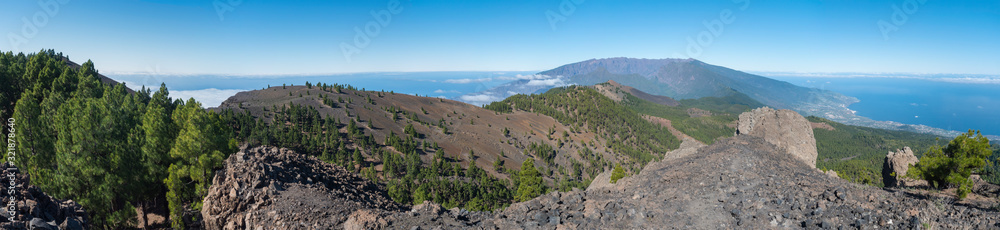 Panoramatic view of volcanic landscape with lush green pine trees, colorful volcanoes and white clouds at path Ruta de los Volcanes, hiking trail. La Palma, Canary Islands, Spain, Blue sky background