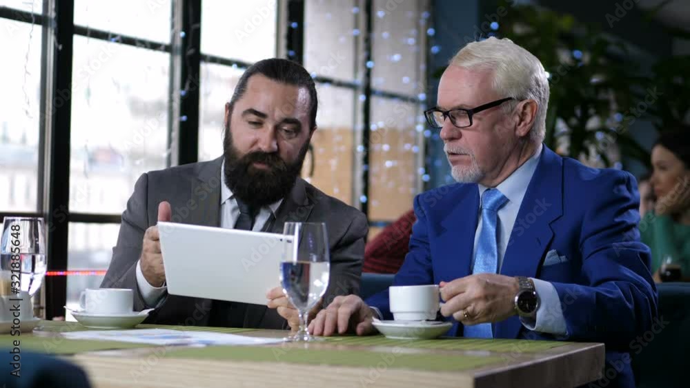 Middle aged man holding tablet showing on charts profit from joint activities to old spectacled white-haired partner with coffee cup. Businessmen in formal clothing during negotiation in cafe Middle aged man holding tablet showing on charts profit from joint activities to old spectacled white-haired partner with coffee cup. Businessmen in formal clothing during negotiation in cafe