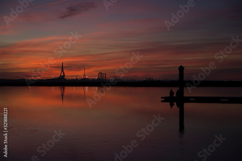 coastal landscape after sunset in blue hour with two people contemplating the horizon on a wooden jetty, with bridge, buildings and lights in the background, reddish and orange colors © roferfrann