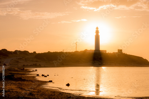 lighthouse with sun from behind at sunset on foot of beach reflected in sea water