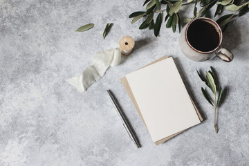 Moody feminine wedding stationery mock-up scene. Blank greeting card, cup of coffee, silver pen with olive leaves, branches. Grunge table background. Flat lay, top view. Mediterranean breakfast.
