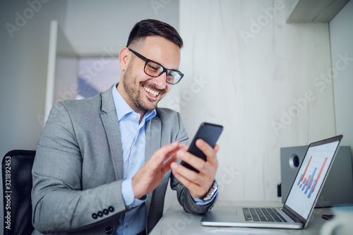 Handsome caucasian classy unshaven businessman in suit and with eyeglasses using smart phone while sitting in office. On table is laptop with chart on it.