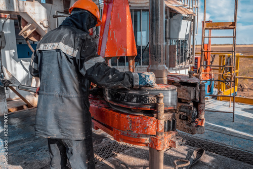 Work driller in red uniform, in helmet and goggles. He uses a hydraulic wrench to screw drill pipes to lower them into an oil well and continue drilling it. The concept of a working person.