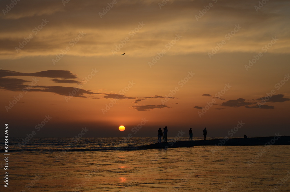 The setting sun over a narrow sand spit with silhouettes of people in the sea