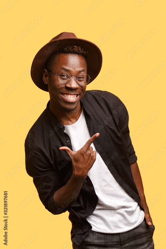 Hilarious African guy in hat, glasses and black shirt poses on a yellow background. Studio shooting. Hilarious African guy in hat, glasses and black shirt poses on a yellow background. Studio shooting.