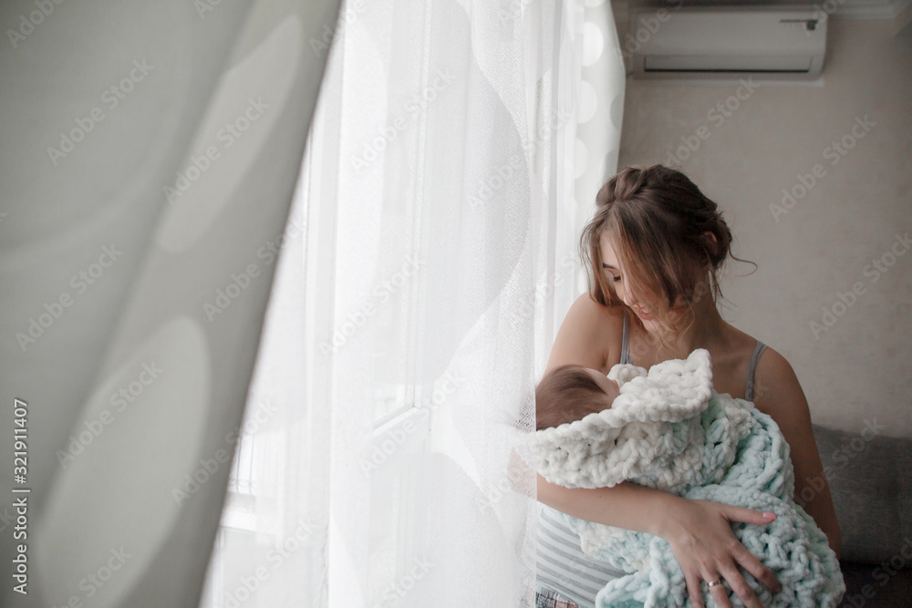 Mother holding the 3 month baby wrapped in a cozy blanket near the window and cradles the infant to sleep Mother holding the 3 month baby wrapped in a cozy blanket near the window and cradles the infant to sleep
