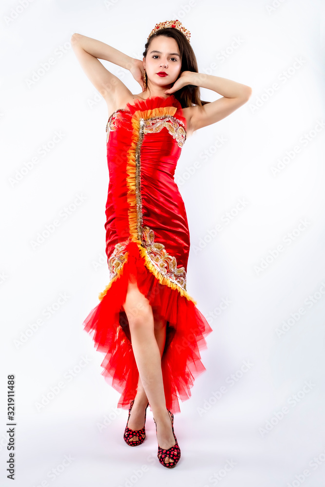 Beautiful brunette adult girl in a red silk chic, luxurious dress wearing gold with red stones crown standing and posing on gray background. Isolate Beautiful brunette adult girl in a red silk chic, luxurious dress wearing gold with red stones crown standing and posing on gray background. Isolate