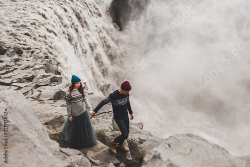 Young stylish couple in love walking together near famous Icelandic Dettifoss waterfall. Traditional wool sweaters, hat, red hair, gray skirt. Dramatic nordic landscape, cold weather in Iceland