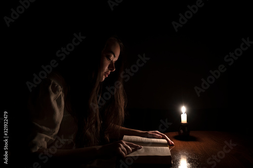 A young girl reading a book at night with candle light. She is wearing an old white dress. Dark background. Side view. 
