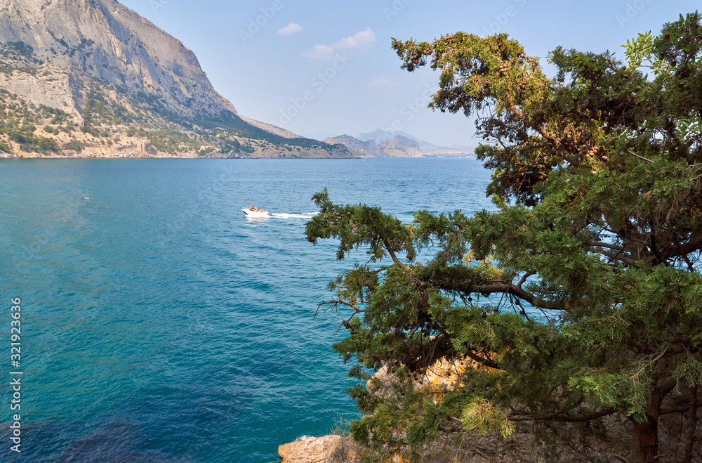 Boat moving on the waves. View of Green Bay, coastal cliffs and a ridge of mountains on the horizon. Mountain juniper. Black Sea coast, Sudak region, Republic of Crimea Boat moving on the waves. View of Green Bay, coastal cliffs and a ridge of mountains on the horizon. Mountain juniper. Black Sea coast, Sudak region, Republic of Crimea