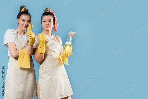 Two happy teenage girls with cleaning equipment in the studio are leaping on blue. Yellow rags and sponges, liquid for washing and dusting. Copy space.