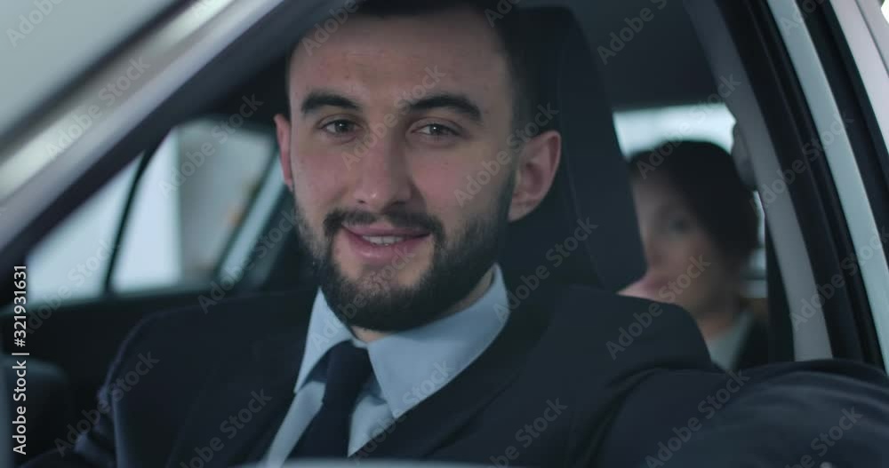 Close-up face of young handsome Caucasian man looking at camera and smiling. Professional chauffeur sitting on driver's seat in car with passenger at the background. Cinema 4k ProRes HQ. Close-up face of young handsome Caucasian man looking at camera and smiling. Professional chauffeur sitting on driver's seat in car with passenger at the background. Cinema 4k ProRes HQ.
