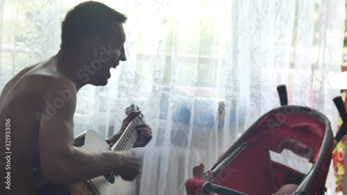 elder man is playing on acoustic guitar and singing song for his little grandson who laying in red baby carriage at window background. Side view medium shot in 4K video.