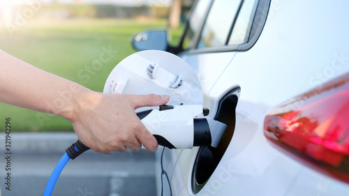 Electric car or ev is charging at station . man use  white power cable and plug on nature background. eco and clean energy concept with the mobile phone smart technology. zero gas and no transmission.