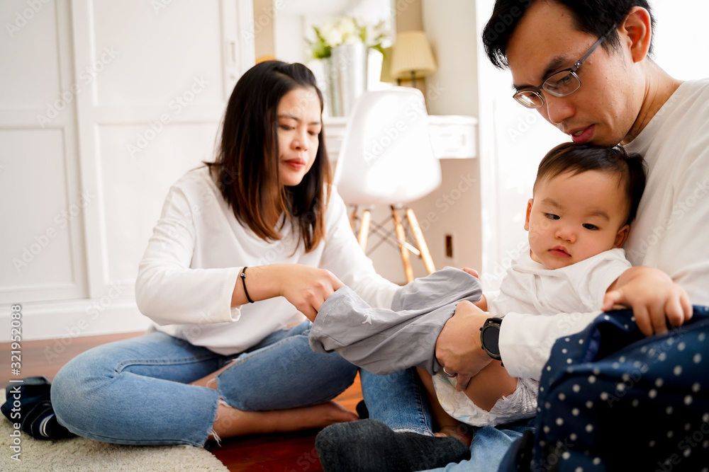 Young Asian father sitting on floor and holding infant boy while mother holding baby clothing and together taking care of son and changing diapers Young Asian father sitting on floor and holding infant boy while mother holding baby clothing and together taking care of son and changing diapers